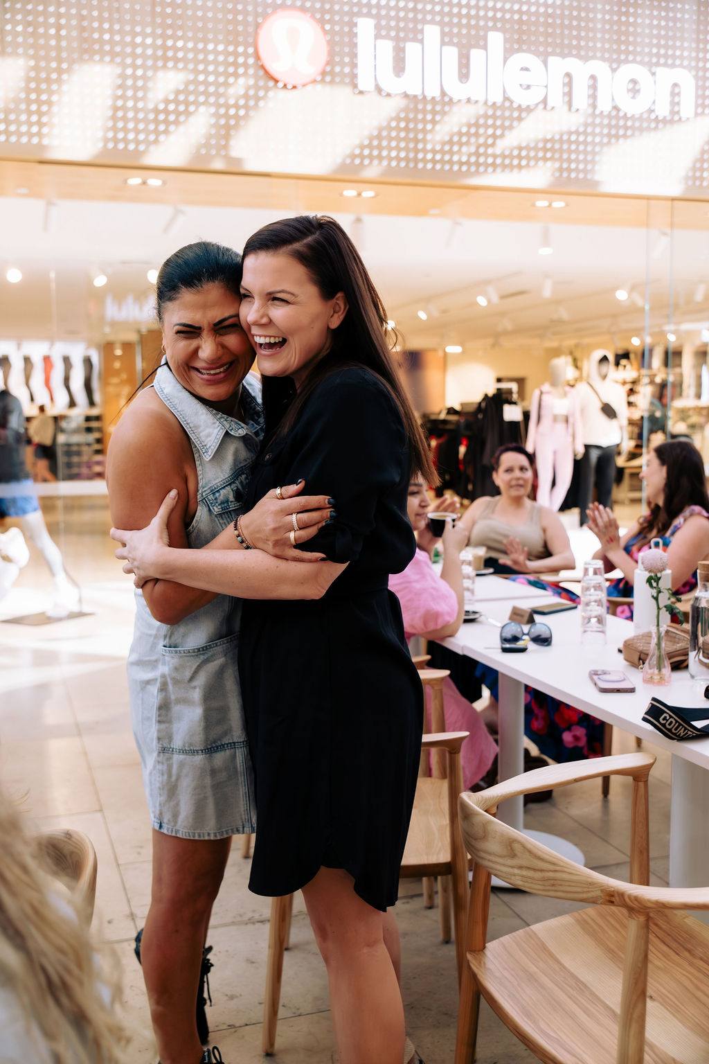 Two women hugging while friends clap at the table behind them